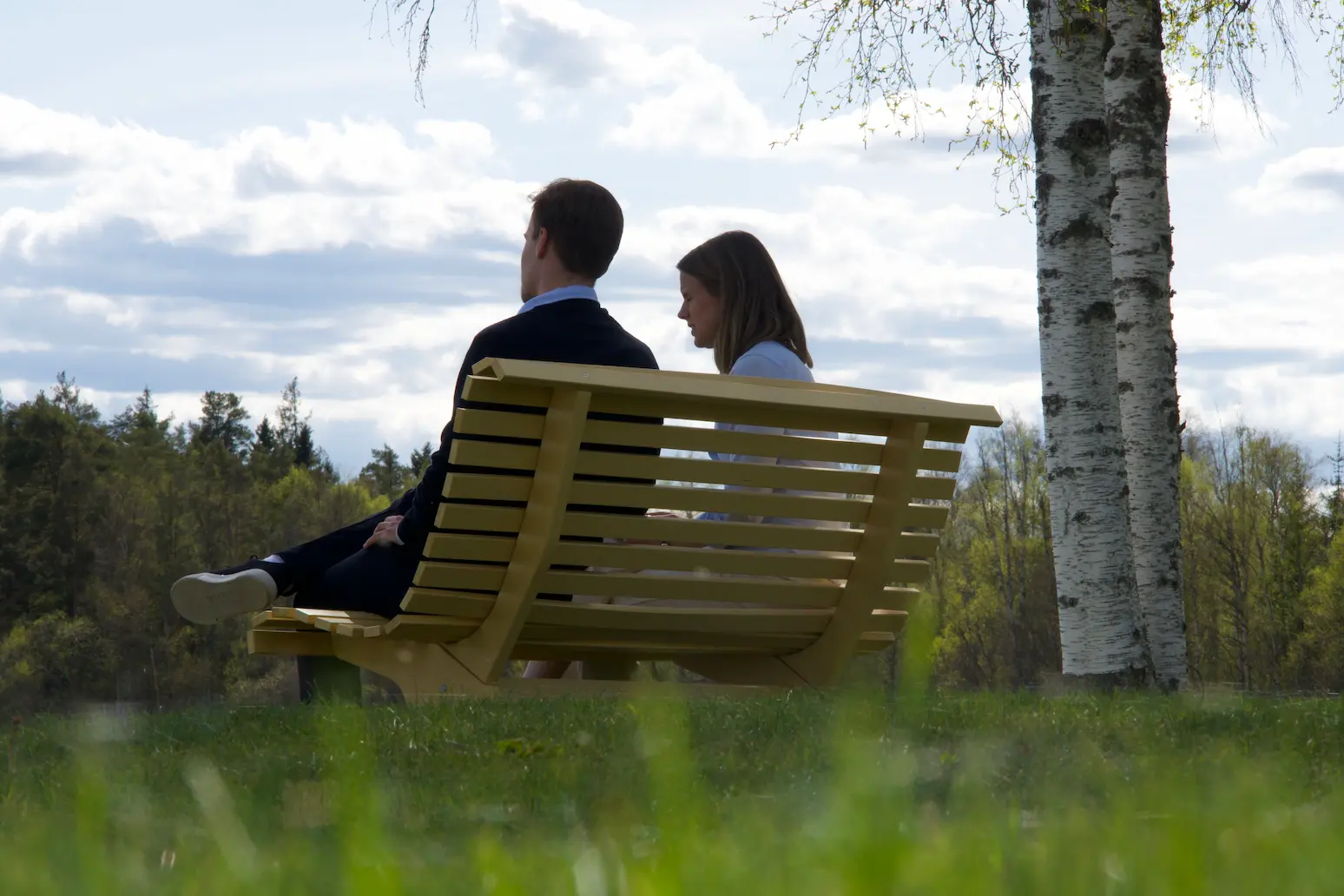 Två personer sitter tillsammans på en gul träbänk vid sjön vid Wiredaholm, omgivna av björkar och grönska